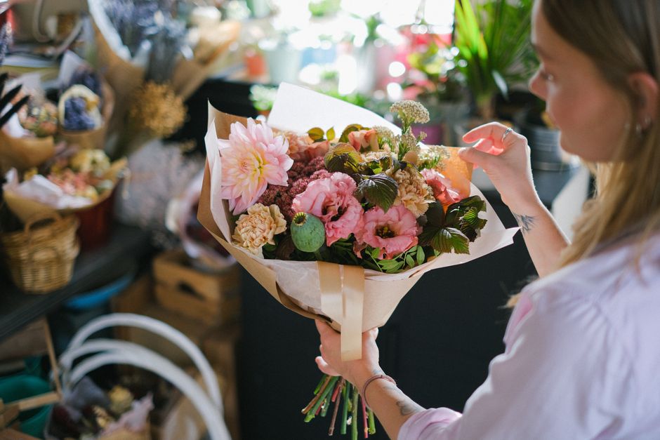 A woman in a light-colored shirt holds a large, vibrant bouquet of fresh flowers in a florist shop. The arrangement includes pink dahlias, light pink lisianthus, beige carnations, and dark green folia