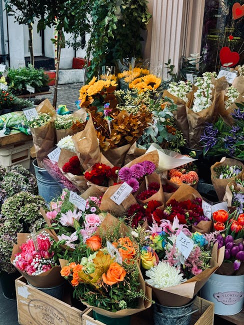A colorful display of fresh flower bouquets arranged outdoors, featuring a variety of blooms including bright yellow sunflowers, deep red roses, pink lilies, purple irises, and white chrysanthemums, e