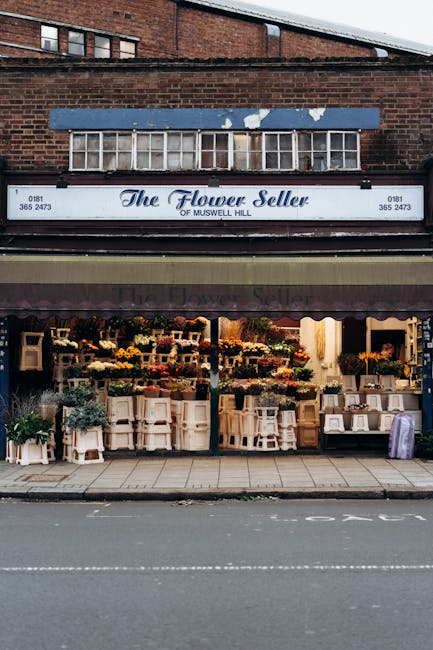 The storefront of The Flower Seller of Muswell Hill displaying a variety of fresh floral arrangements on white wooden shelves and tables. The display includes vibrant bouquets with yellow, pink, purpl