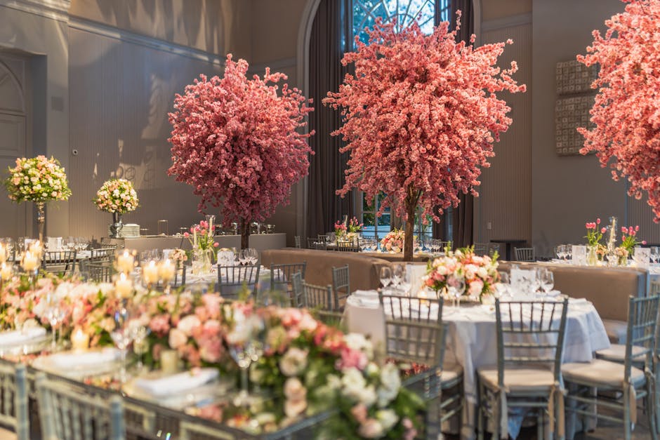 An elegantly decorated banquet hall featuring large, vibrant pink cherry blossom trees as the centerpiece arrangements, surrounded by round tables draped with white tablecloths. Each table is adorned 