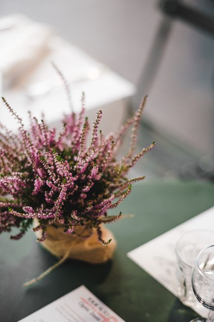A lush bouquet of pink and purple heather flowers arranged in a small, rustic wrapping at the center of a dark, smooth table. The arrangement features delicate, elongated stems with tiny blossoms dens